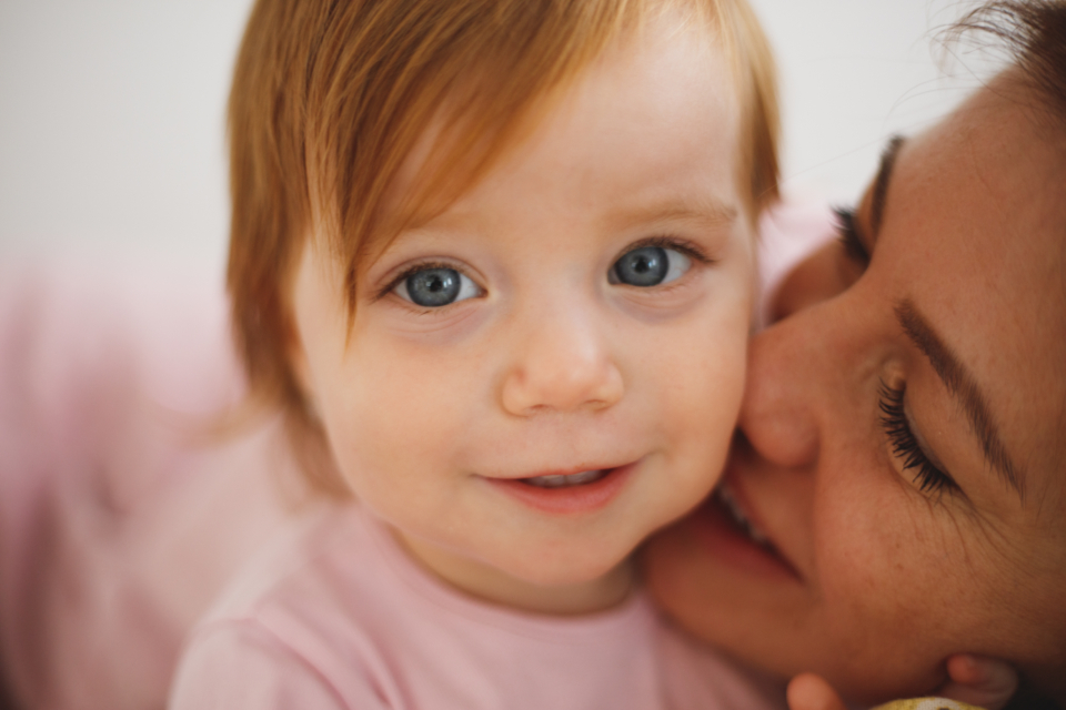 A mother kissing her baby on the cheek while the baby looks at the camera