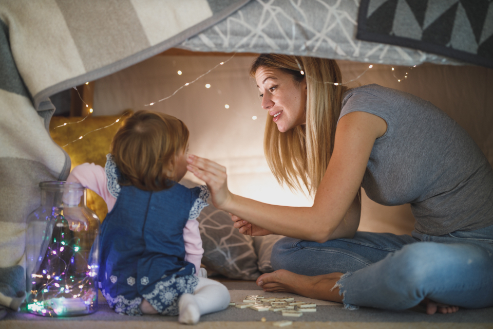 A mother sitting with her toddler inside a blanket fort strung with fairy lights